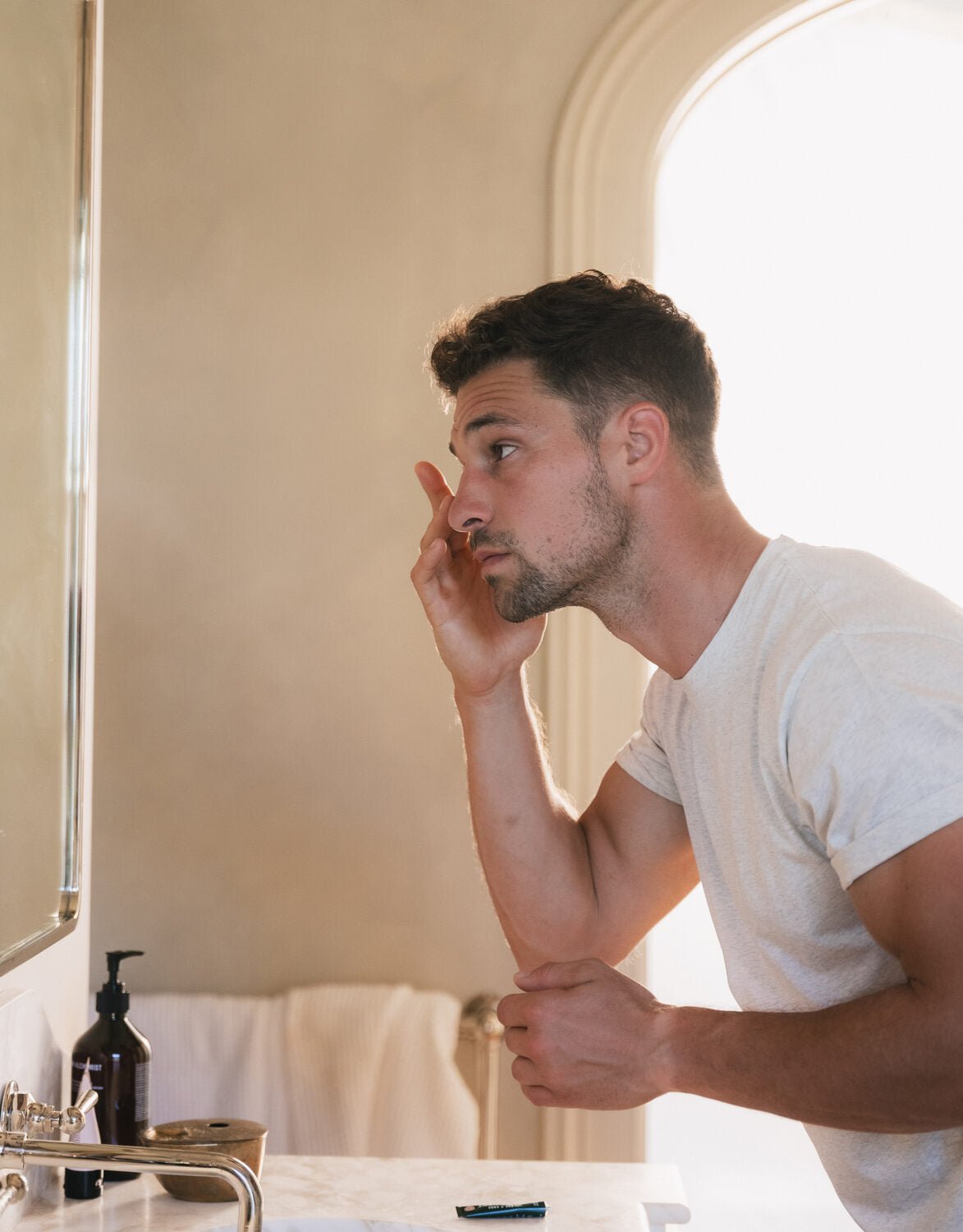 A man leaning into a bathroom mirror in morning light, working eye serum into the skin around his eye