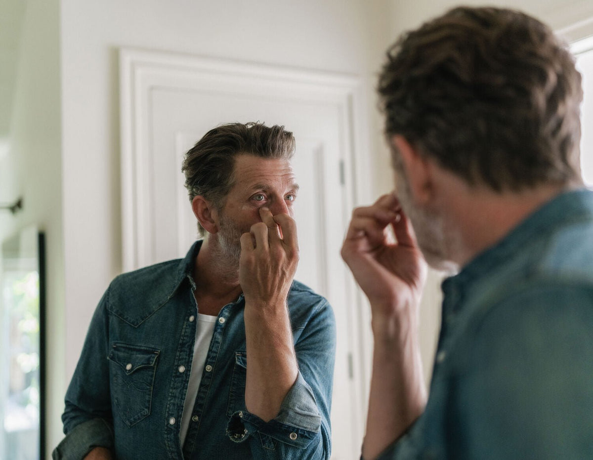 A man applying eye serum at a mirror, his reflection beside him