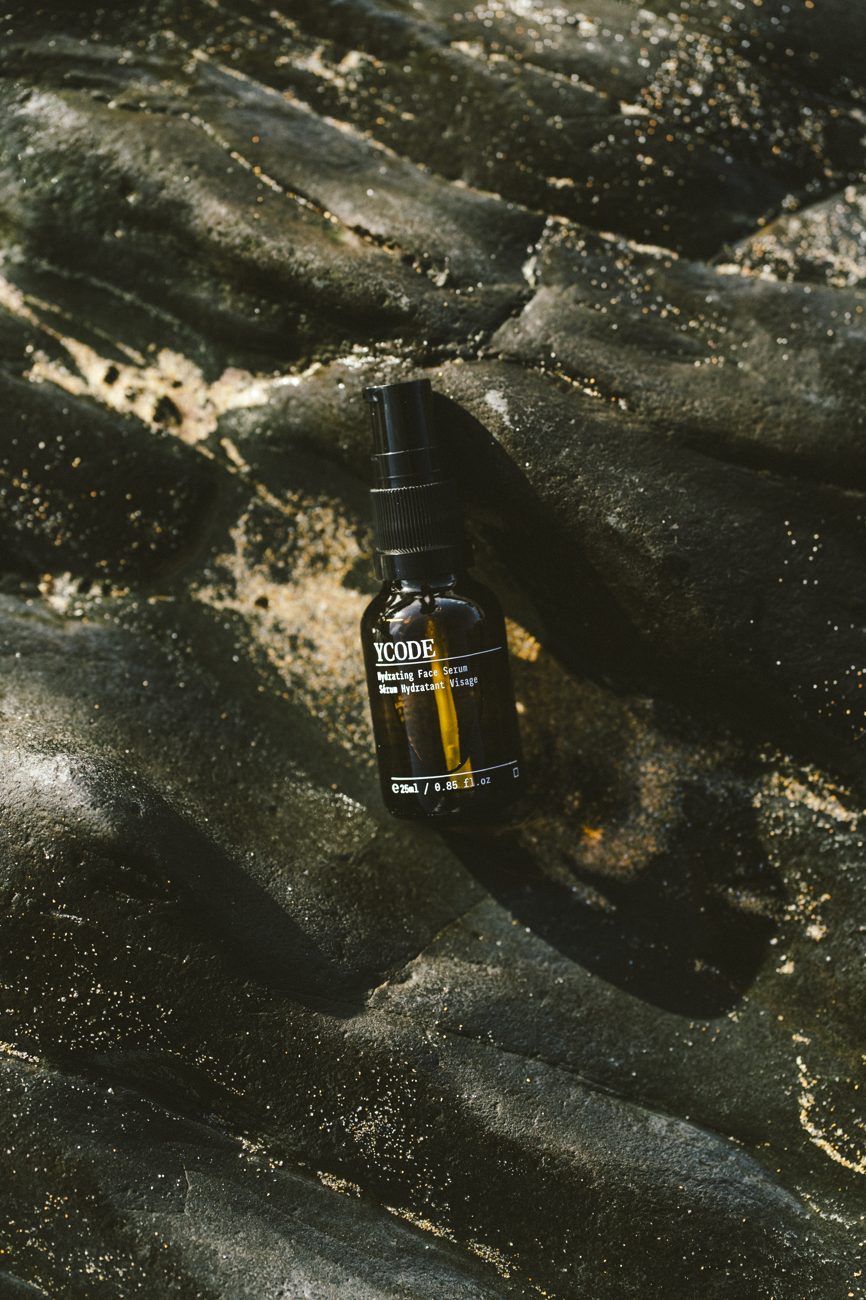 A YCODE serum bottle resting on wet black sea rock in low sun